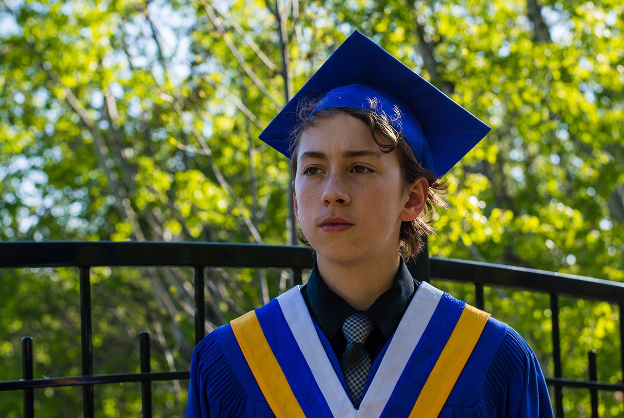 Young man in graduation cap and gown looks ahead with determination against a backdrop of a black wrought iron fence and lush green trees