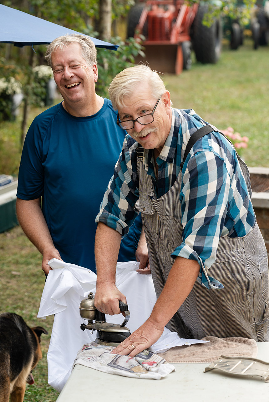 Best man, dressed in coveralls, uses an antique steam iron to prep his dress shirt outdoors at the countryside wedding venue
