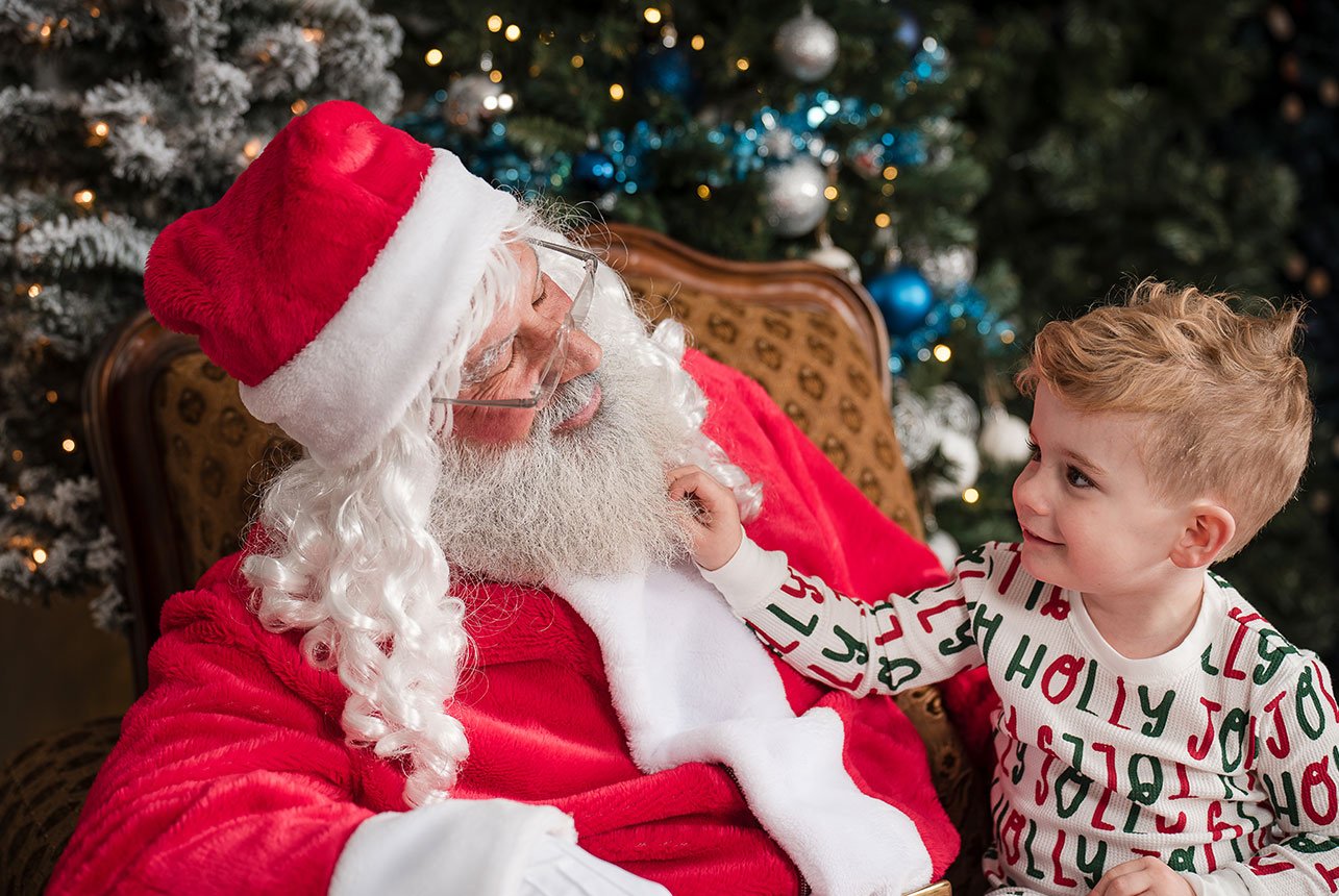 Boy sits on Santa’s knee, smiling as he gently tugs Santa’s beard during a cozy Christmas mini session at Catriona Hope Photography’s studio in Sherwood Park.