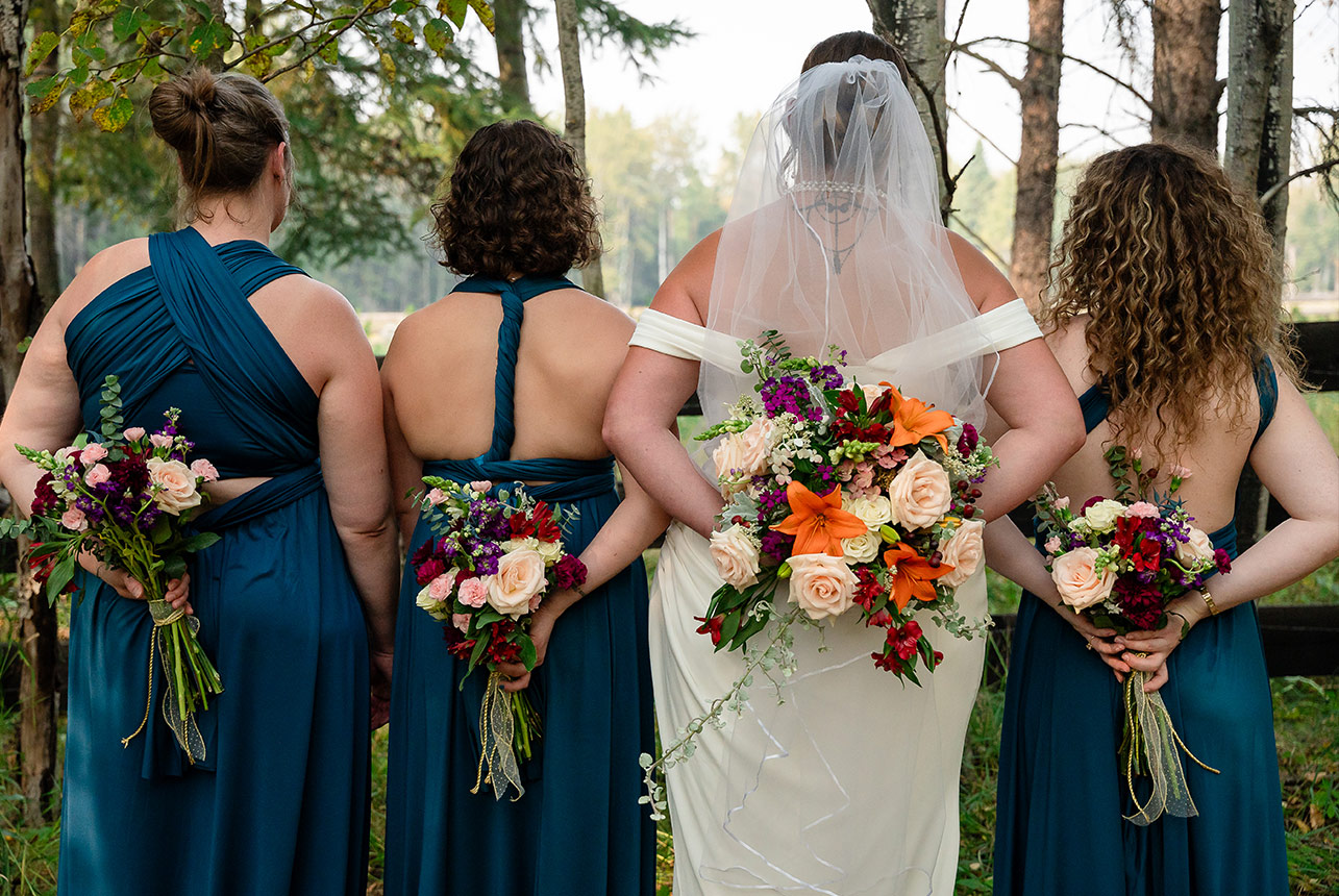 Bride and her bridesmaids stand facing away, holding bouquets, their dresses flowing against the countryside backdrop