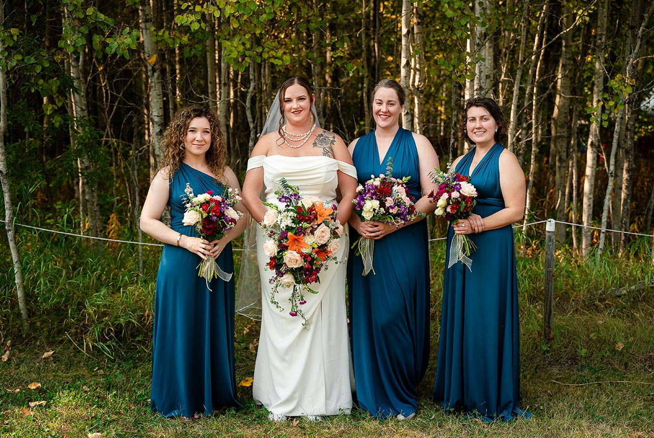 Bride and bridesmaids pose in full-length gowns, holding their floral bouquets in a radiant summer wedding setting