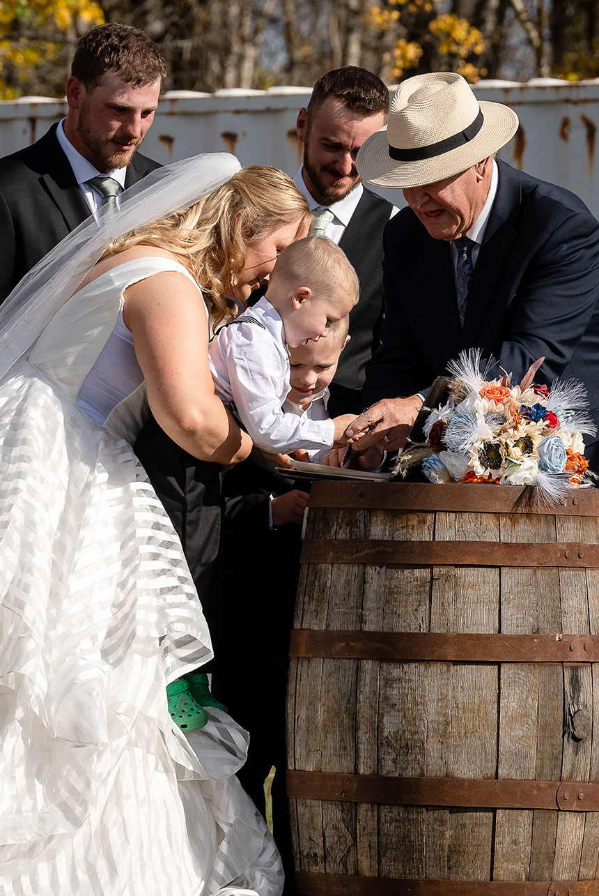 Bride holds her children while signing papers on a rustic wooden barrel, guided by the commissioner, with the groom and best man nearby