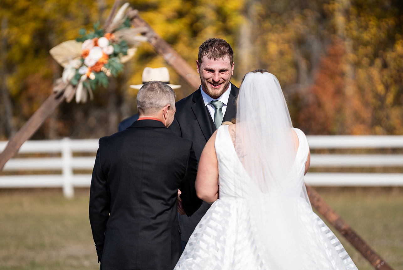 Bride and father share a hidden handshake with the groom under a floral triangle wedding arch at Finnegan Farms