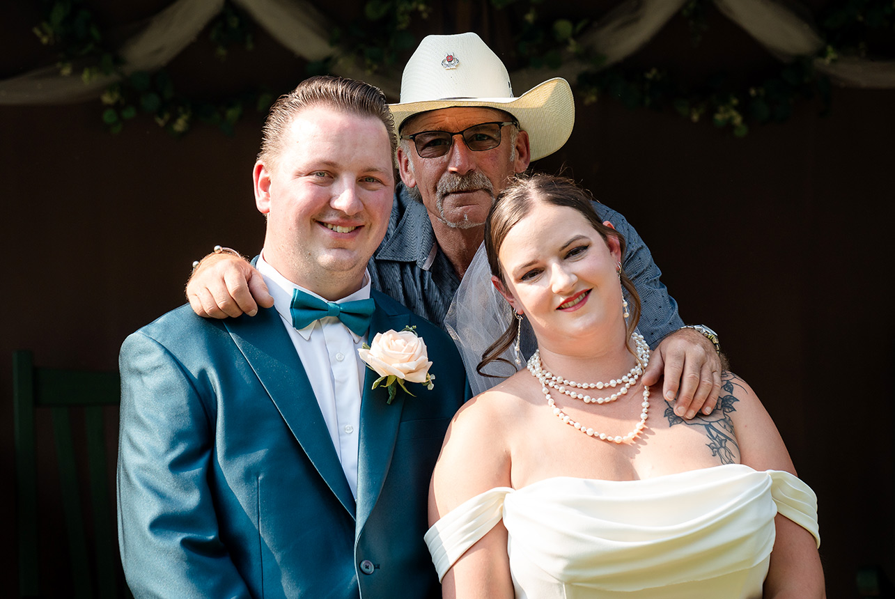 Bride and groom pose with a cowboy guest who stands behind them, resting his arms on their shoulders, sharing a lighthearted moment