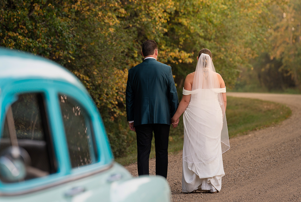 Newlyweds walk hand in hand down a winding country gravel road, surrounded by trees with early autumn leaves beginning to turn