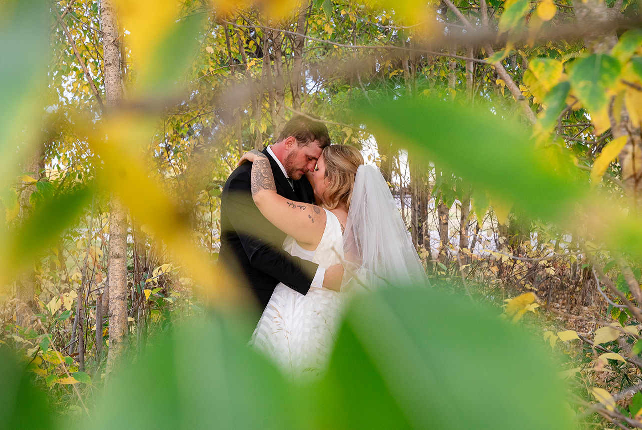 Bride and groom hug under autumn foliage, seen through blurred branches for a magical touch