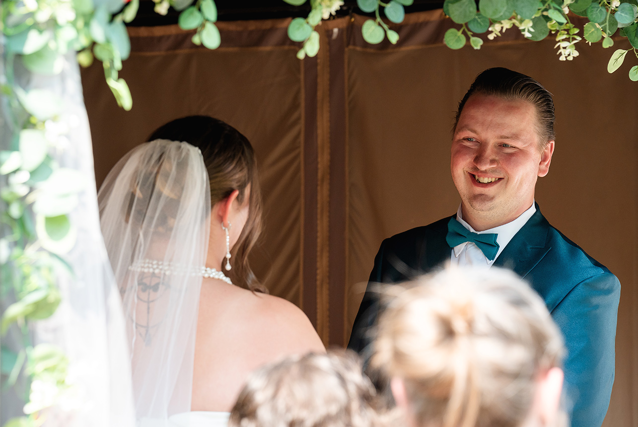 Bride seen from behind as she faces her groom, who smiles warmly under the summer sun during their outdoor wedding ceremony
