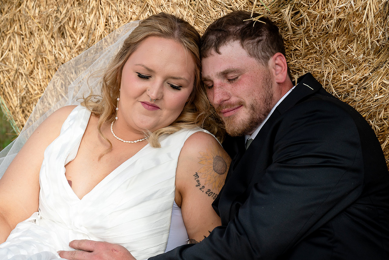 Bride and groom snuggled together, leaning against a haybale on a crisp autumn day, sharing a quiet and intimate moment after their wedding ceremony