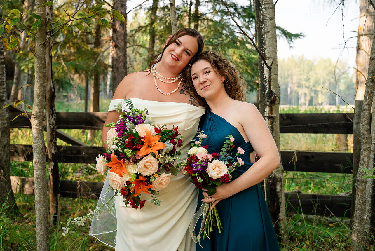 Bride and maid of honor stand near a rustic country fence, surrounded by trees, with an open field stretching into the distance