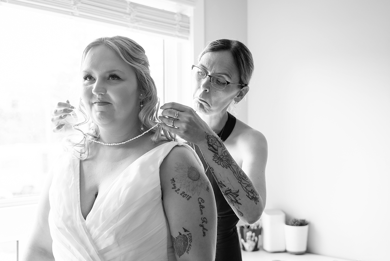 Bride tears up while her mother places a necklace around her, both emotional in a bright room with a window behind them