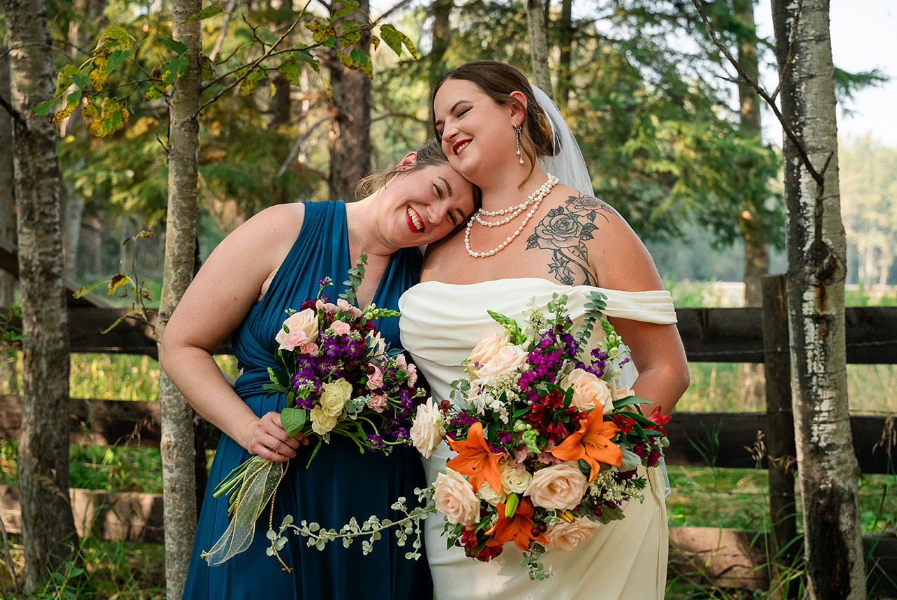 Bride and her sister, a bridesmaid, share a tender moment as she rests her head on the bride’s shoulder, surrounded by trees and countryside