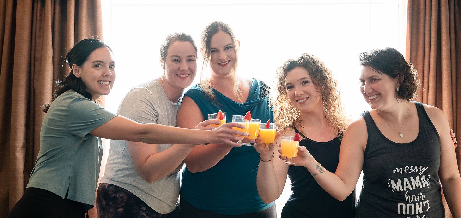 Bridesmaids raise their glasses in a celebratory toast near a bright window, preparing for the wedding day in Westlock, AB