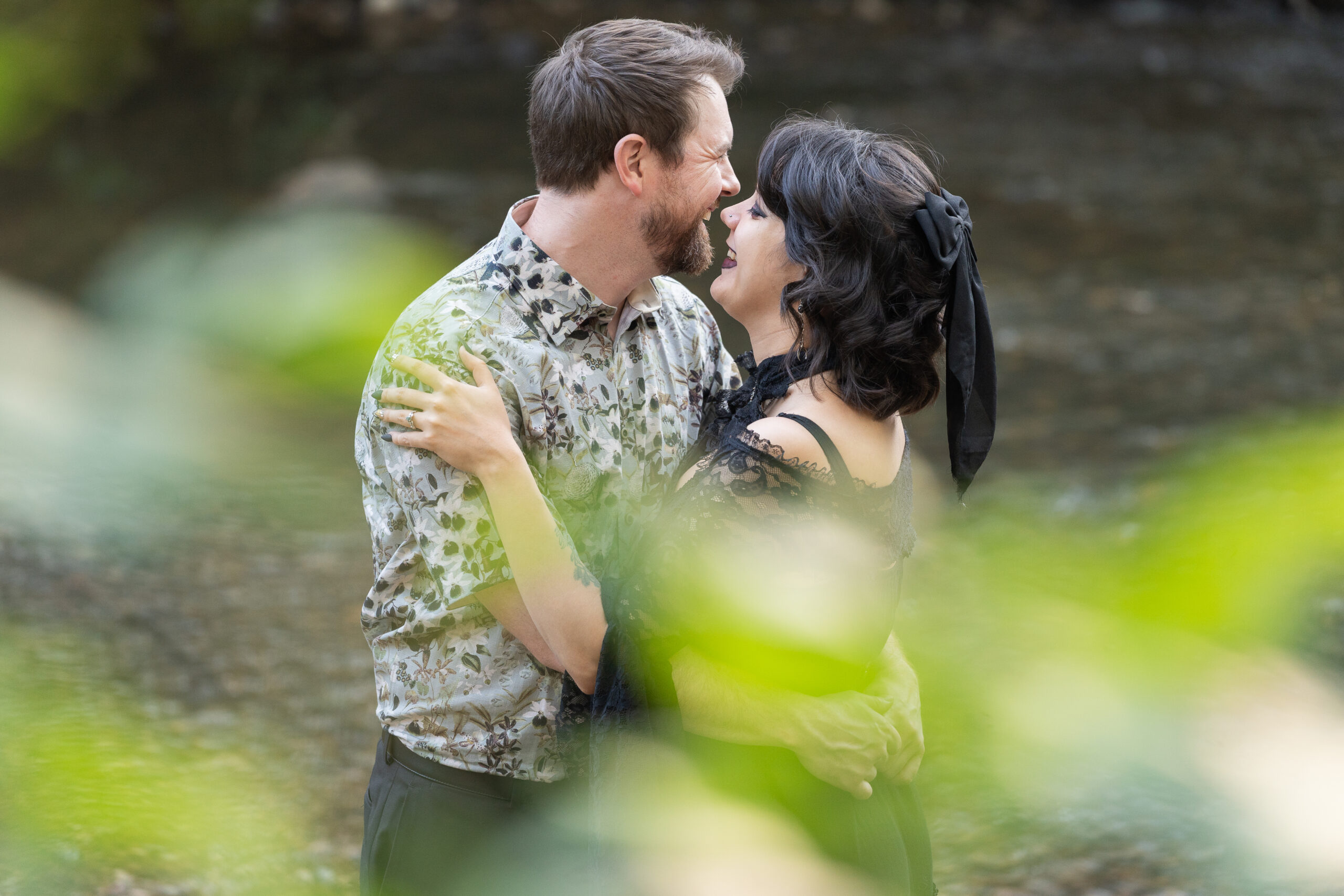 Couple laugh together in a close embrace by the water, framed by soft green leaves