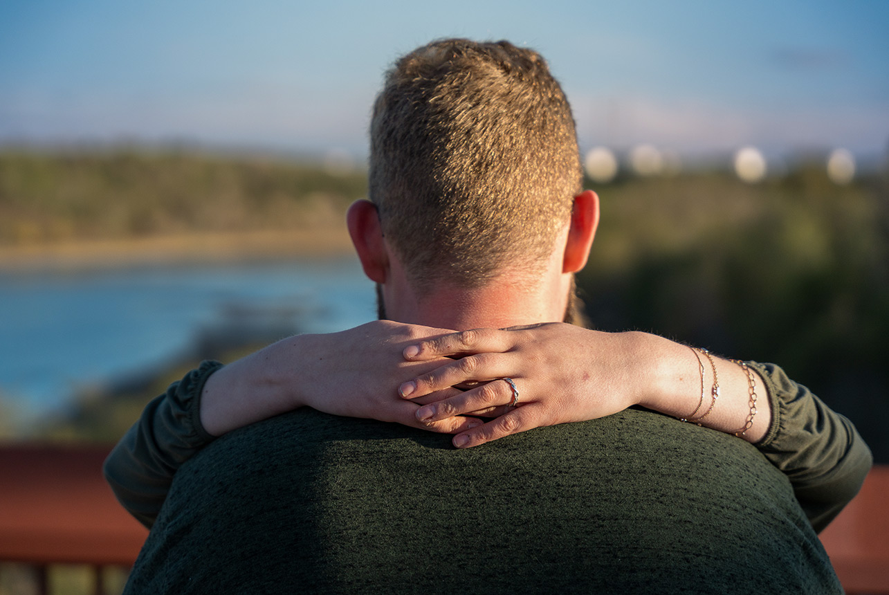 close hands wrapped ring detail back hug Woman’s hands wrapped around her fiancé’s neck from behind, engagement ring glinting in evening light