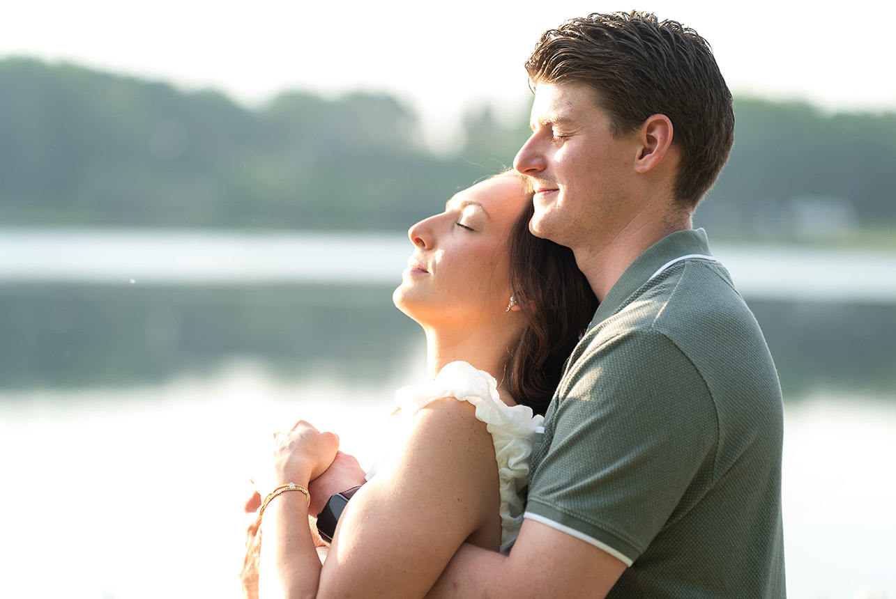 couple arm in arm summer lake portrait Engaged couple standing closely together by the lake, smiling softly at the camera in evening light