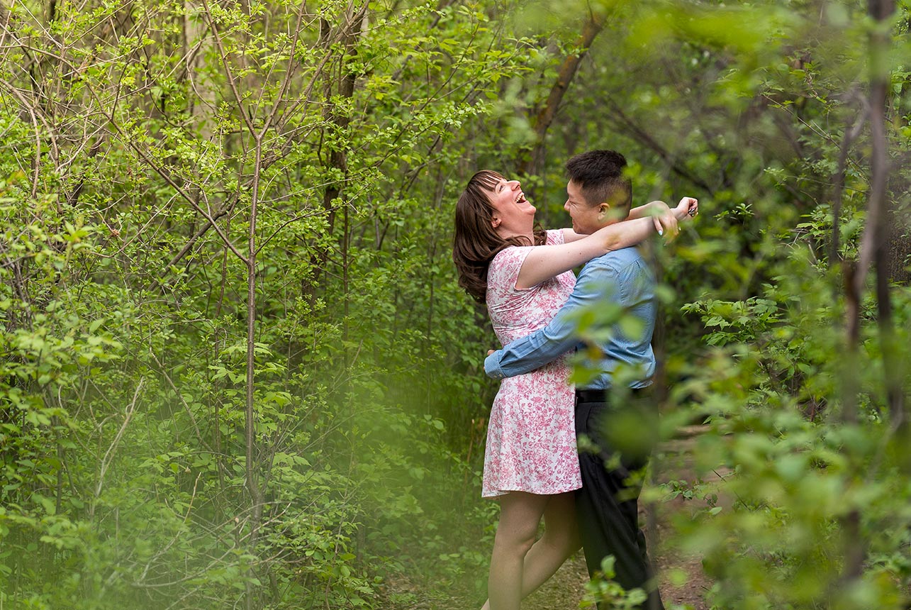 Couple laughing and embracing in a lush green forest during a spring engagement session in Edmonton