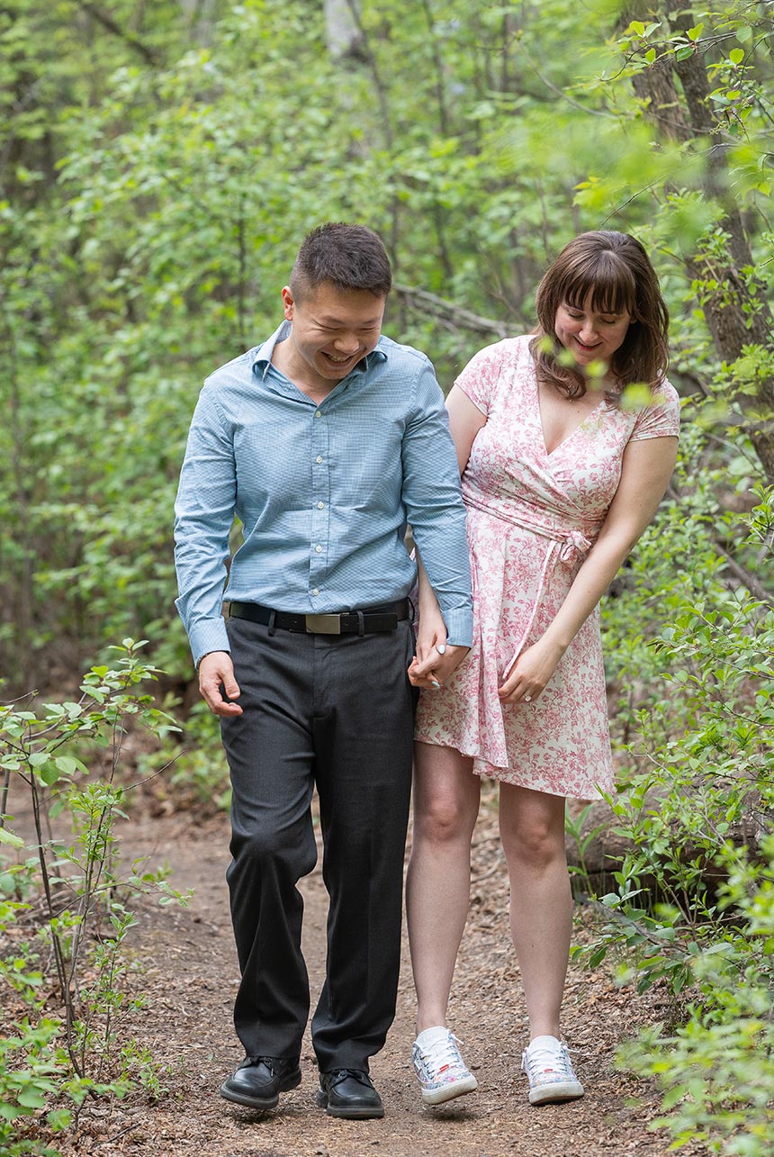 Couple laughing walking spring trail edmonton Couple laughing and walking hand in hand down a quiet forest trail during spring engagement photography in Edmonton