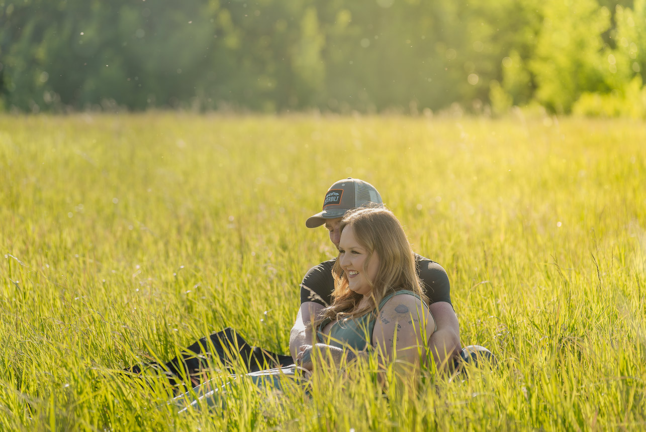 Couple sitting in tall golden grass, laughing together with sunlight dancing through the summer air