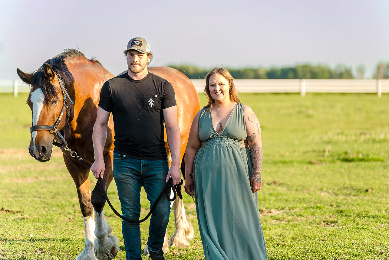 Couple standing with a majestic horse on a grassy field at Finnegan Farms in Redwater during a golden sunset