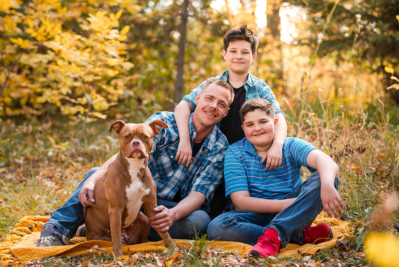 Dad and his young boys sitting on a blanket with their dog in an autumn forest.