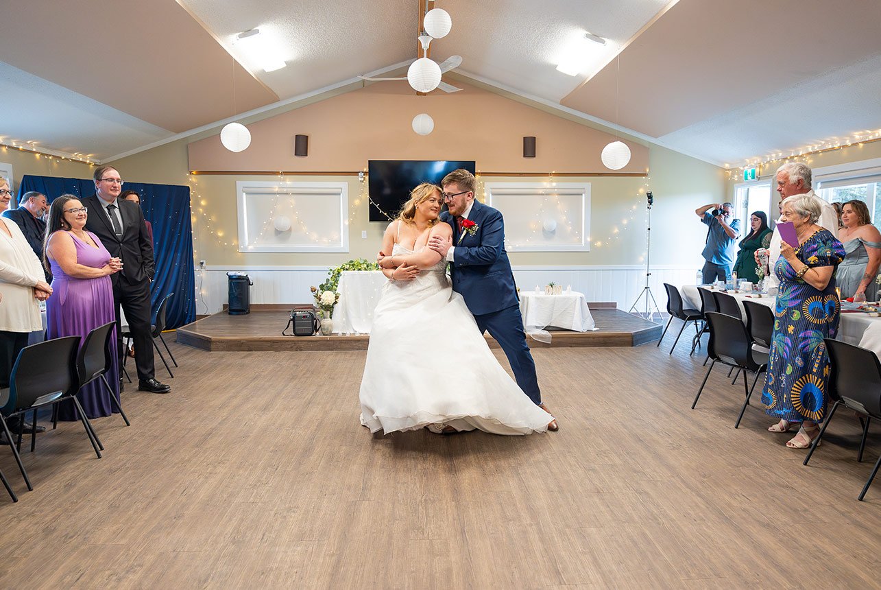 Bride and groom share a playful first dance in a warmly lit Edmonton hall while family and friends watch and smile