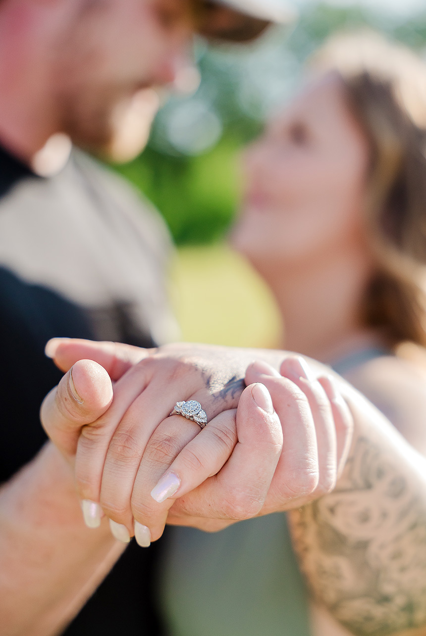 Close-up of engaged couple’s hands with the ring in focus and their faces softly blurred in the background