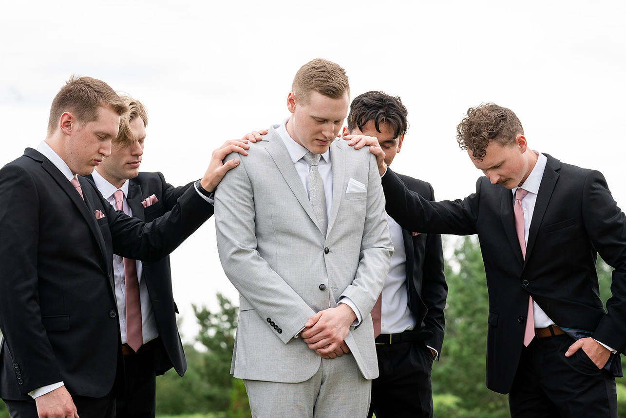 Groom surrounded by groomsmen in prayer with hands laid on his shoulders at Trinity Baptist Church
