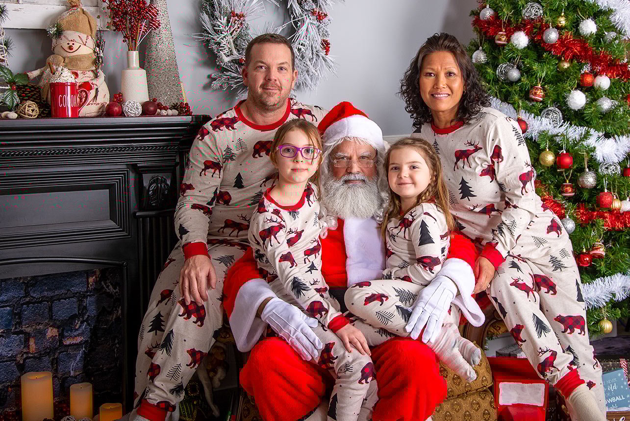 Family of four sitting on Santa's chair with Santa in the middle, all in pajamas, with a fireplace and Christmas decor on the side.