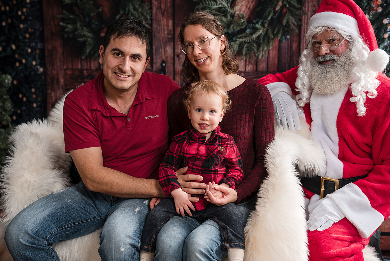 Parents and toddler smile with Santa beside them on a fur-draped seat in a festive setting
