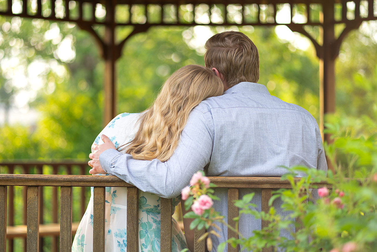 Couple sit on a wooden gazebo bench with backs to camera; she rests her head on his shoulder while he wraps an arm around her