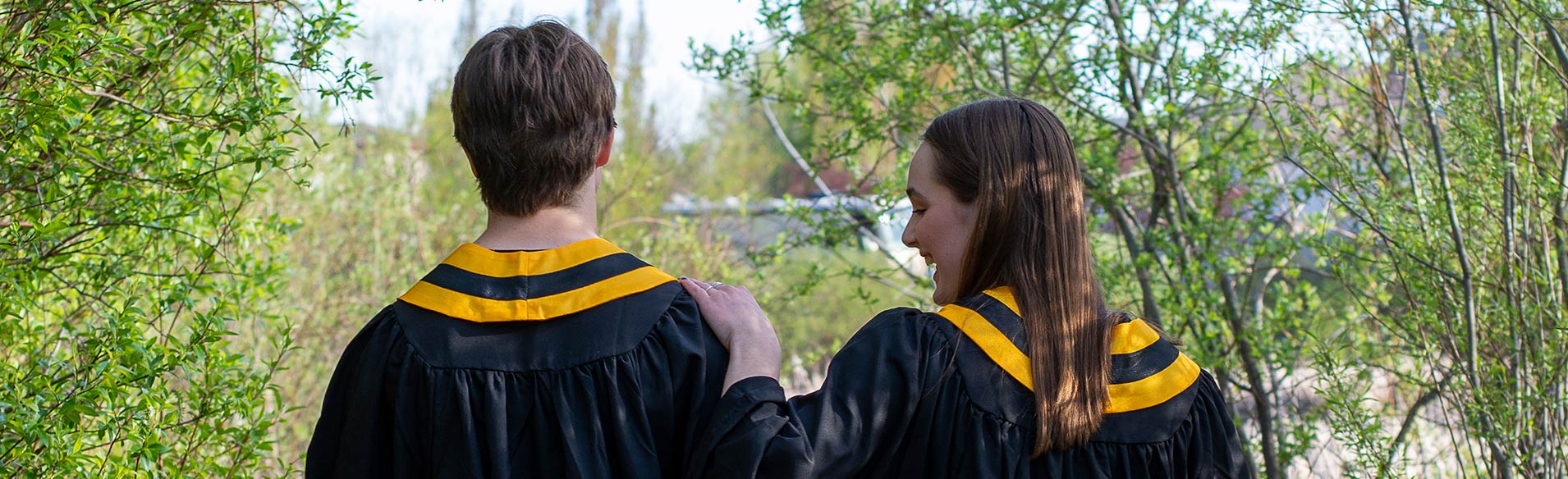High school graduates, a boy and girl, in graduation gowns with backs to camera, laughing and celebrating on sunny path surrounded by trees.