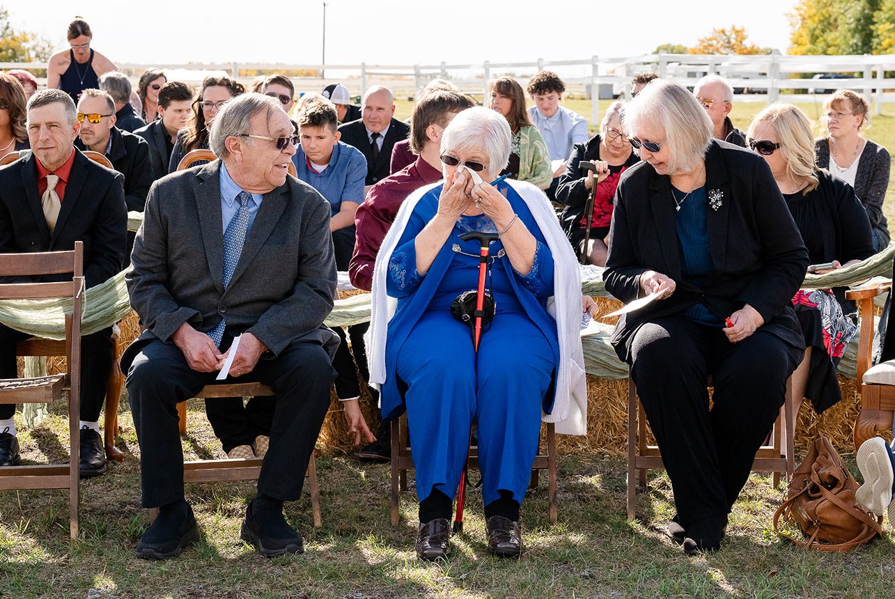 Great-grandmother wipes away tears during the wedding ceremony, seated in the front row with guests behind her
