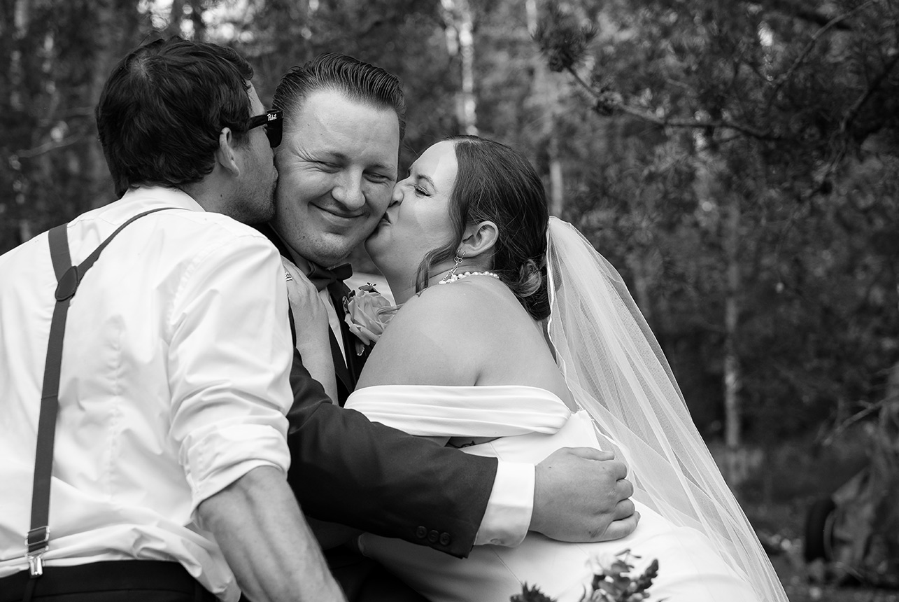 Groom winces with a playful cringe as he gets a kiss on one cheek from his bride and on the other from a groomsman during a fun wedding moment