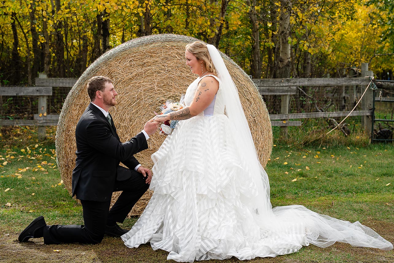 Groom on one knee holding the bride’s hand as she looks at him against a rustic farm background