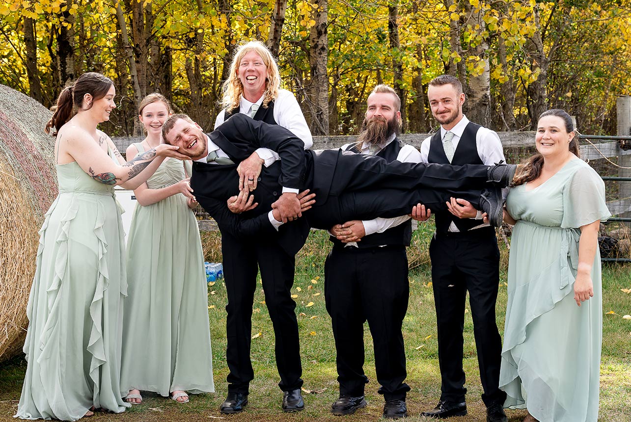 Groomsmen and bridesmaids hold the groom up in a playful group photo with haybales and a rustic farm background