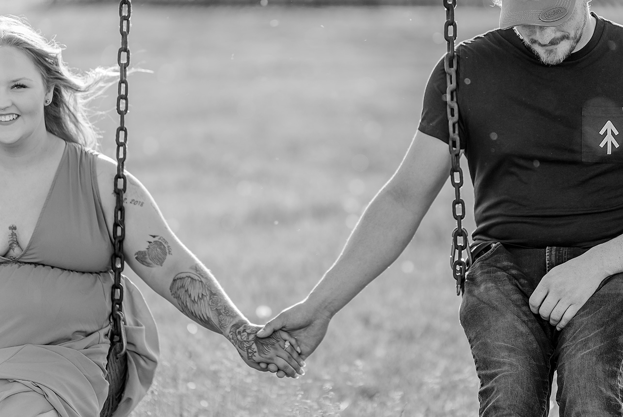 Black and white photo of couple on swings holding hands, her tattooed arm reaching out in love