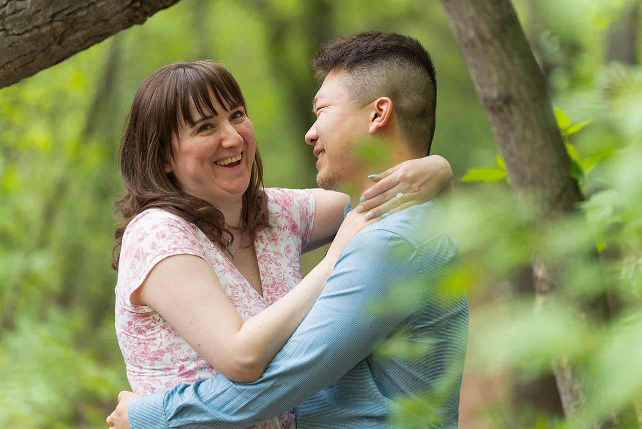 joyful couple spring forest candid smile Engaged couple sharing a laugh surrounded by trees during a spring session in Edmonton’s River Valley