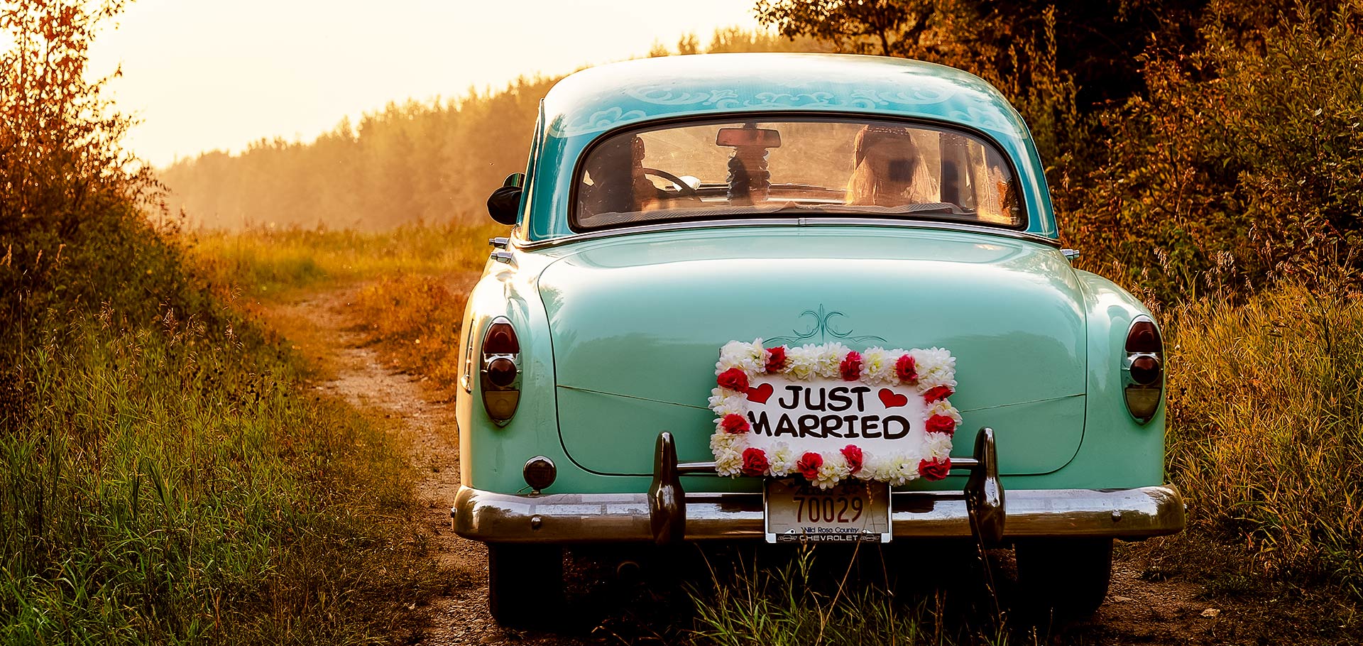 Bride and groom drive down a country road at sunset, a ‘Just Married’ sign on the back of their car as they head into the open fields