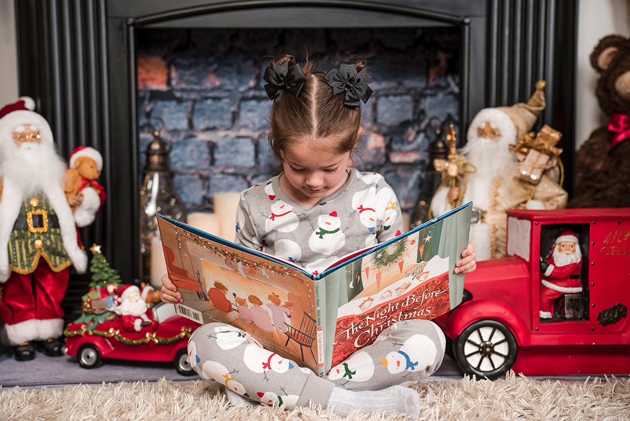 Little girl in pajamas, sitting cross-legged, reading 'The Night Before Christmas' book in front of a fireplace surrounded by Christmas decor.