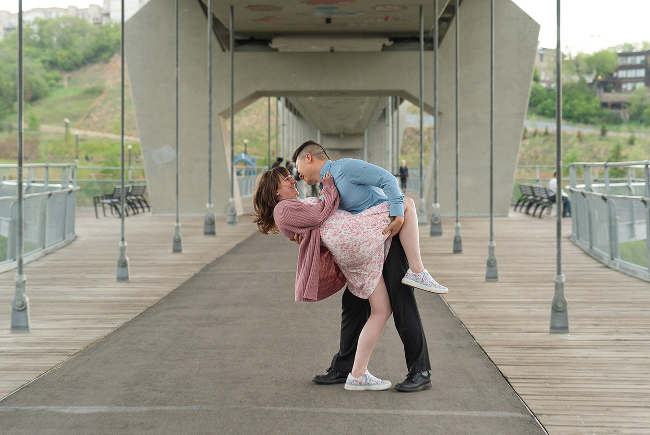 playful dip kiss under edmonton bridge Engaged couple laughing as he dips her under a pedestrian bridge in Edmonton during their spring engagement session