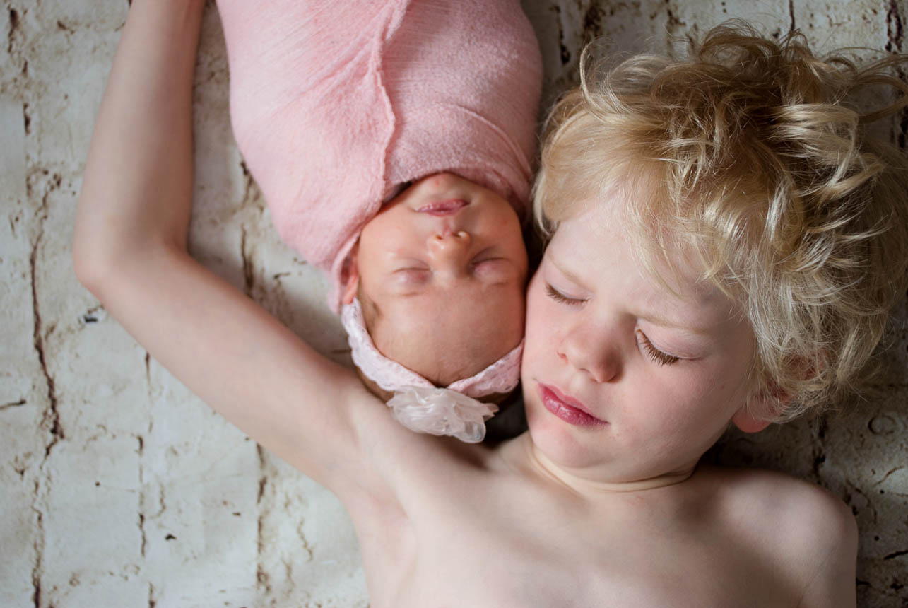 A young brother and his newborn sister, lying back to back, captured in a studio portrait.