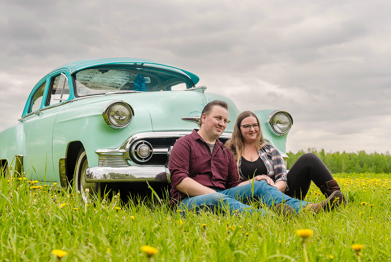 Couple sit in a meadow, leaning against a vintage teal car and smiling
