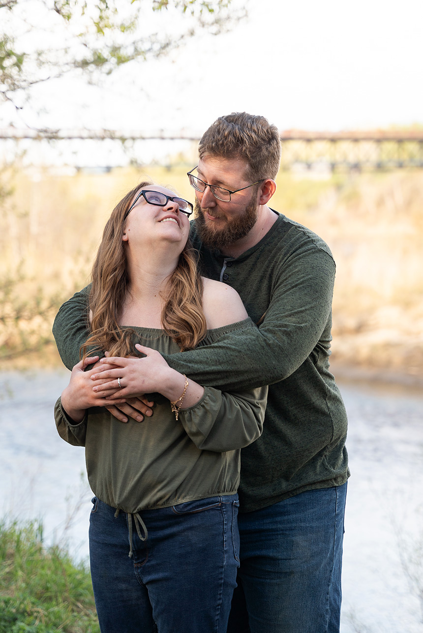 Engaged couple sharing a heartfelt embrace and smile near the riverbank during spring in Edmonton’s Rundle Park