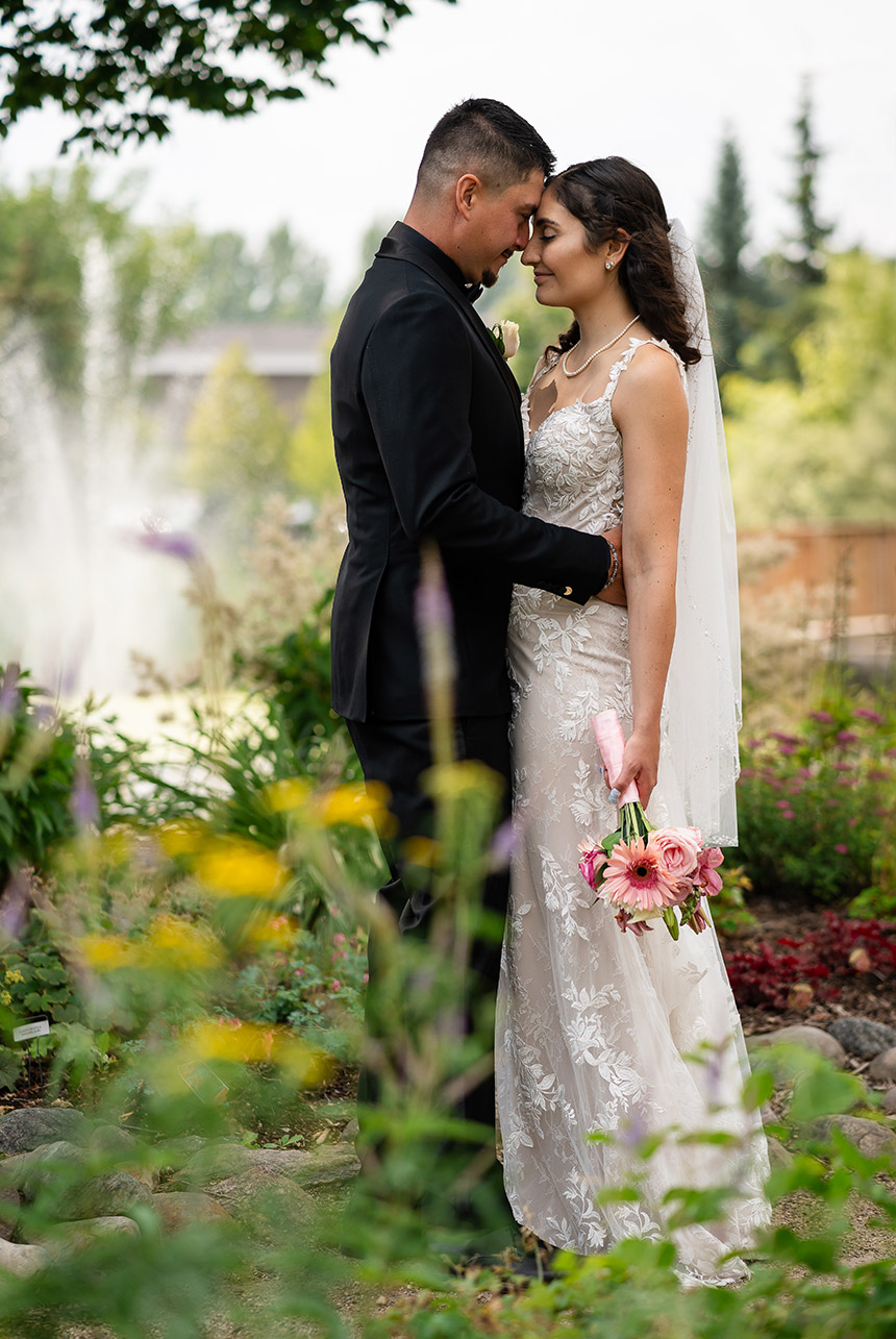 Bride and groom share a quiet forehead-to-forehead moment among flowers, bouquet of pink gerberas in hand, with a soft fountain behind at Greenland Garden Centre in Sherwood Park