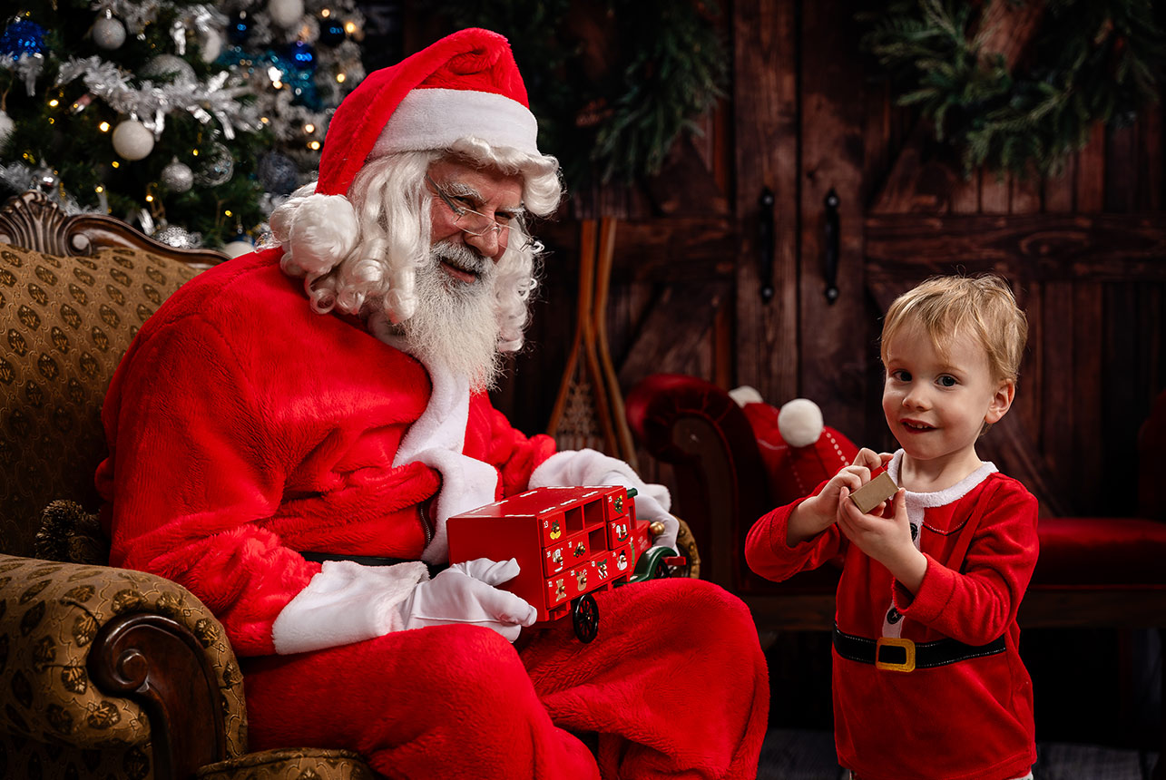 Santa smiles while a young child explores a red toy truck with tiny drawers in a festive studio