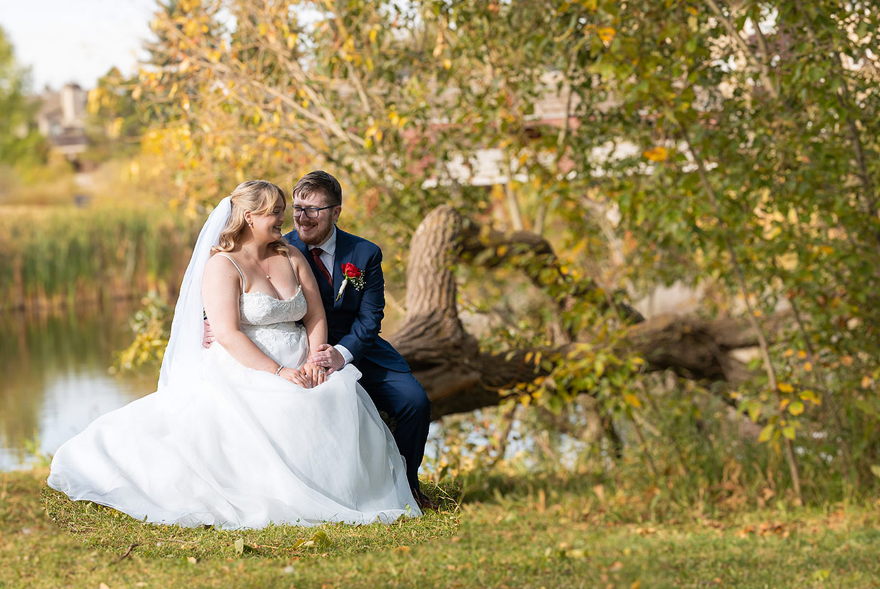 Bride and groom seated by the pond at Woodbridge Farms in Sherwood Park, sharing a smile under golden autumn leaves—white gown, navy suit, red rose boutonniere