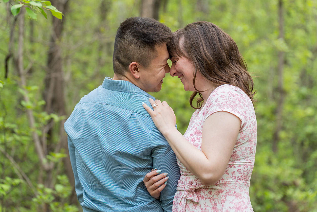 Engaged couple touching foreheads and smiling tenderly in a forest clearing in Edmonton’s River Valley during spring
