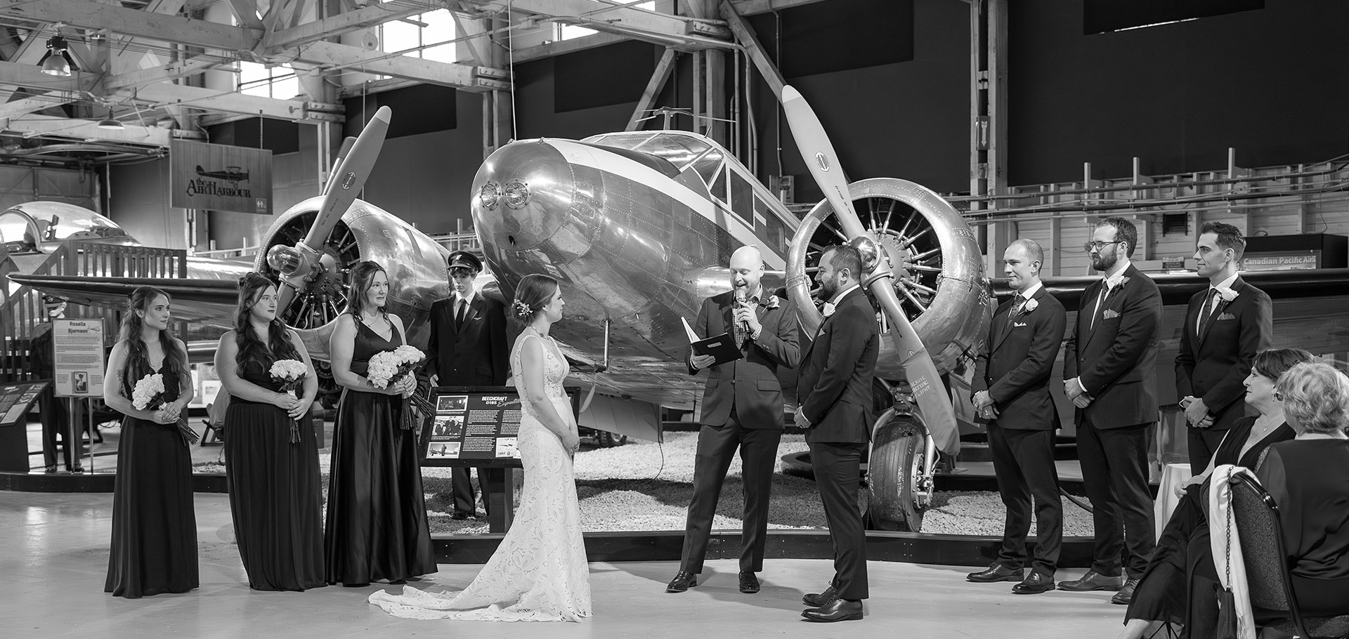 Bride and groom face the officiant during a black-and-white ceremony inside an aviation museum, with a polished vintage aircraft behind them
