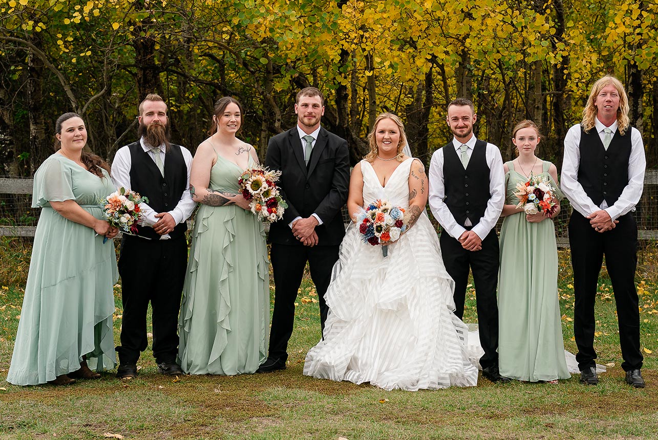 Wedding party posing by a rustic fence with autumn trees arching above them in a natural canopy