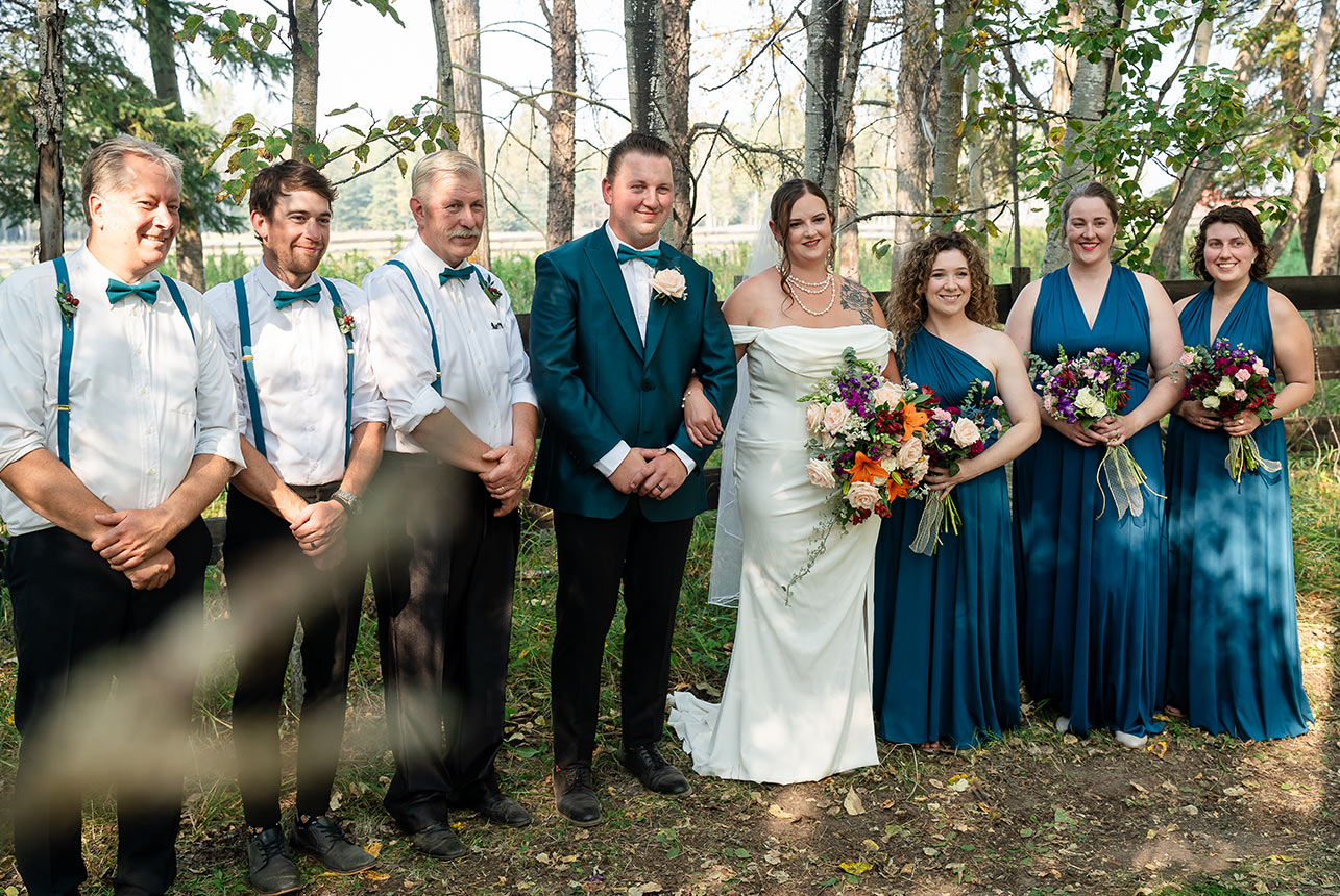 The full wedding party lines up along a dirt road, with a rustic wooden fence and trees creating the perfect countryside backdrop